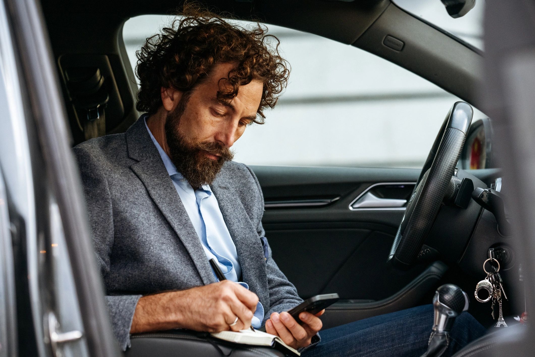 Commuter in a car using a phone and notebook with coffee nearby